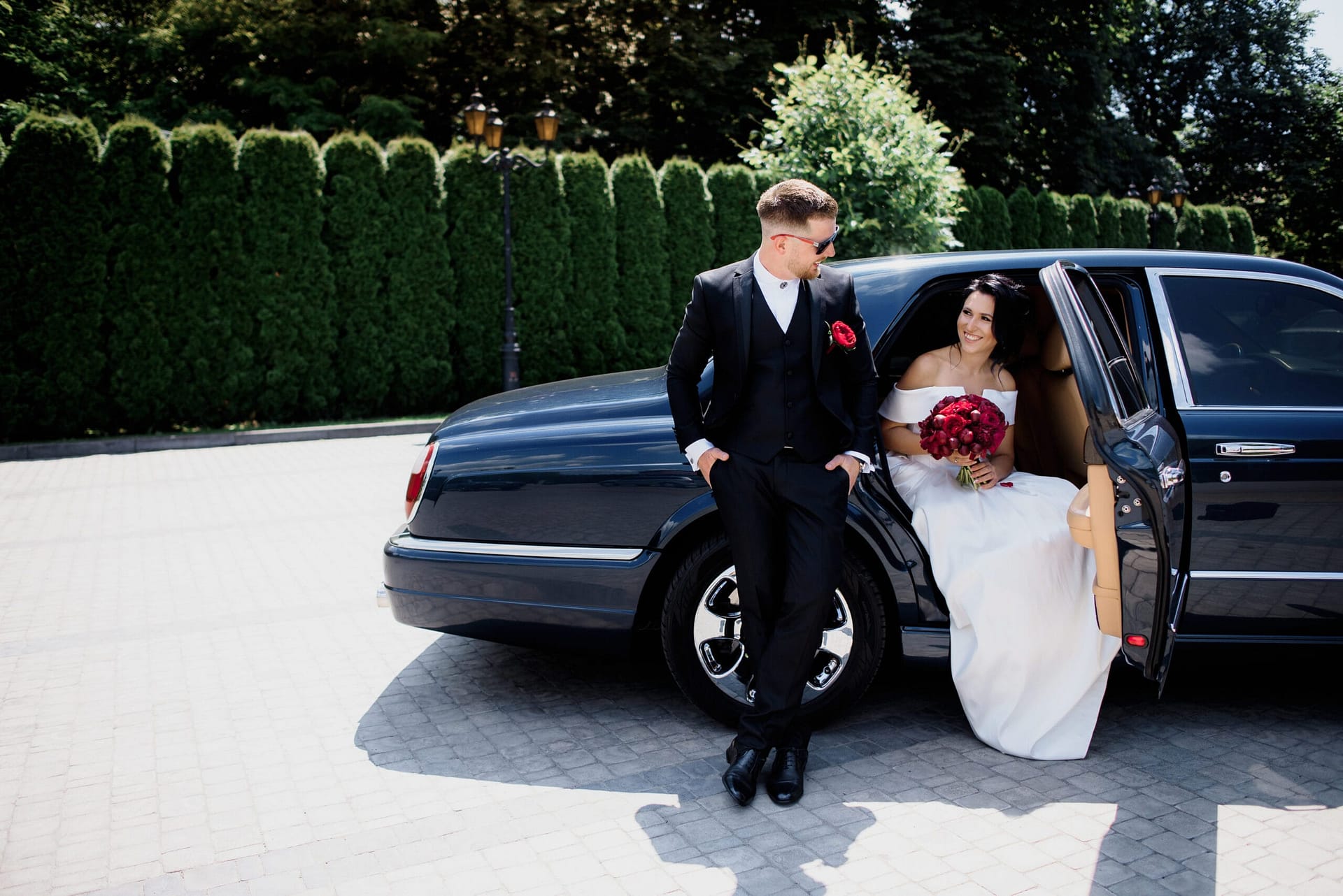beautiful wedding couple is smiling black car sunny day dressed elegant wedding outfits with red bouquet scaled